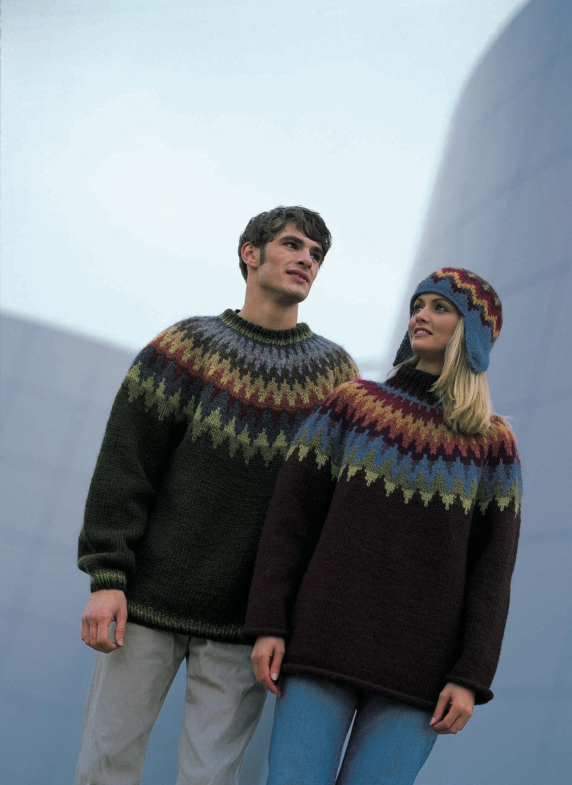 A young man and a woman by a curved metal building, in the pattern’s original colours, most no longer made. He wears Design 2 — very dark green with light green spikes, a slate band, rust, a khaki zigzag, dark brown and flecked grey bands, and striped ribs. She wears Design 1 — dark red-brown with rolled edges, a yoke from light green spikes through blue, plum, rust and gold to wine red, and a tall neck not given in the leaflet — and the ear-flap hat. His trousers and her jeans are not part of the pattern