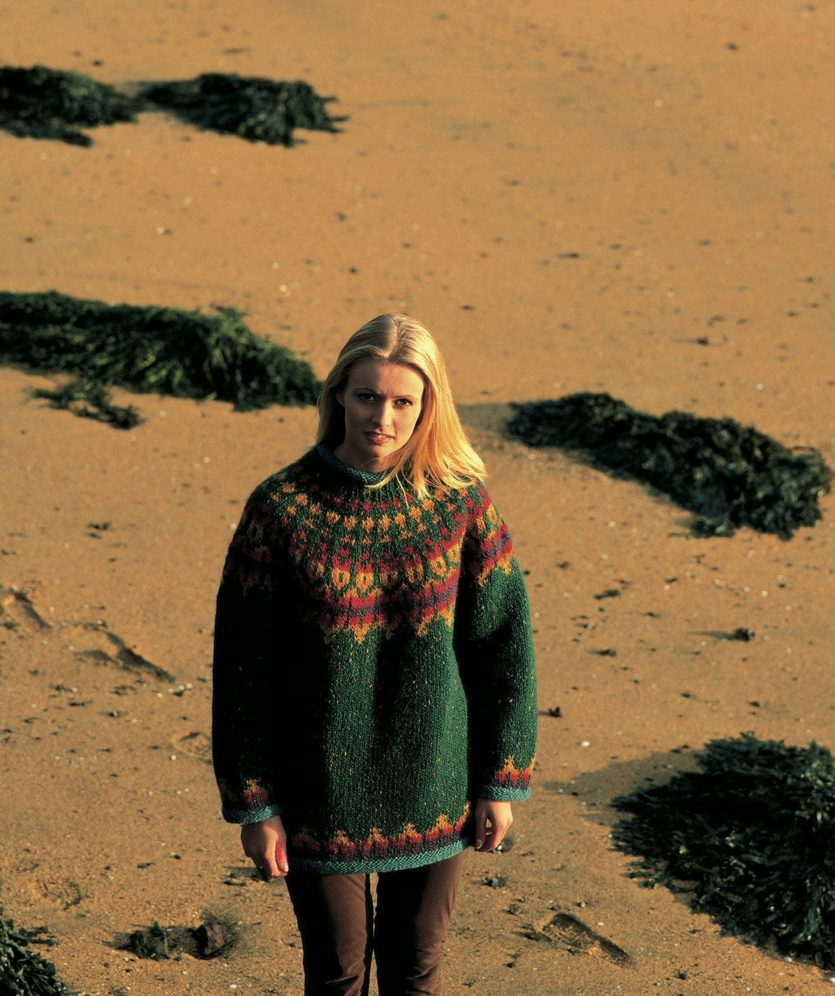 Woman on a beach in warm low sun in the Lopi 18 no. 11 sweater — loose, in dark green tweed, with a yoke of gold diamonds, a band of orange, red and purple flames and gold tulip shapes, the same flames above the hem and cuffs, and teal rolled edges (shown in the original colours — this kit has a plain, lighter garden green with no flecks, a more muted golden heather, a redder amber heather, a darker happy red and a much lighter, bluer arctic exposure). The brown trousers are not part of the pattern