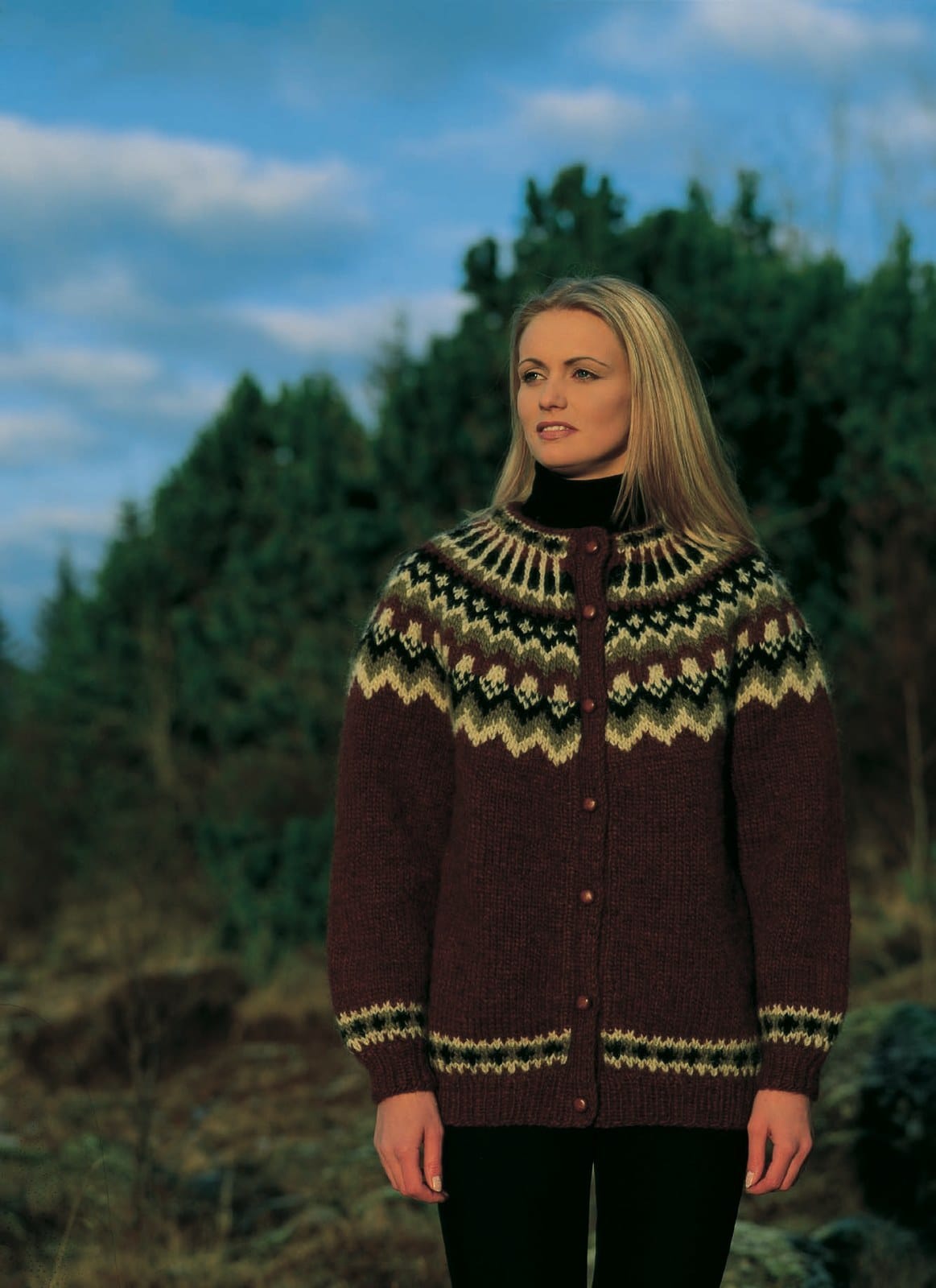 Woman by a stand of trees in the Lopi 18 no. 15 cardigan, buttoned — a dark red-brown body, a yoke of pale zigzags and small pale motifs on olive green and black with pale spokes at the neck, and the same small border above the hem and cuffs (shown in the original colours, in warm low sun — this kit has chocolate heather, less red, ray of light and highland green for 9118, 9718 and 9240). The black polo neck and trousers are not part of the pattern