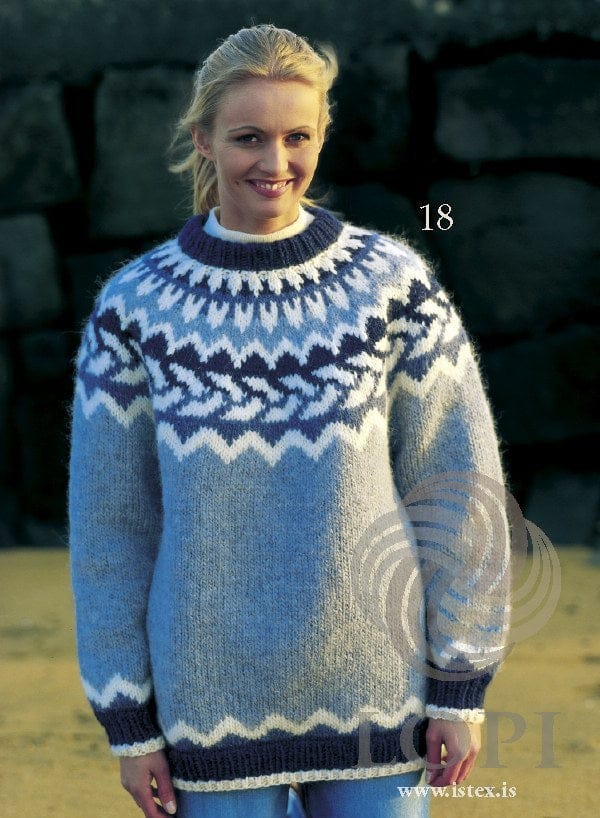 Woman by a stone wall in the Lopi 18 no. 18 sweater — light denim blue with a yoke of a white zigzag, white slanting leaves on dark denim, a navy band and white spikes up to the folded navy neckband, and a white zigzag above the navy hem and cuffs; the white polo neck and the jeans are not part of the pattern
