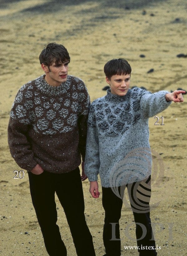 Two young men on a beach. On the right, marked 21 and pointing, the Lopi 18 no. 21 sweater — light grey tweed with a yoke of raised, dark marled cells and a rib collar (shown in the original colours, in dull light — this kit has 9974 light grey tweed, a paler grey, for the original 9451). On the left, marked 20, the brown version, sold as a separate kit; the black trousers are not part of the pattern