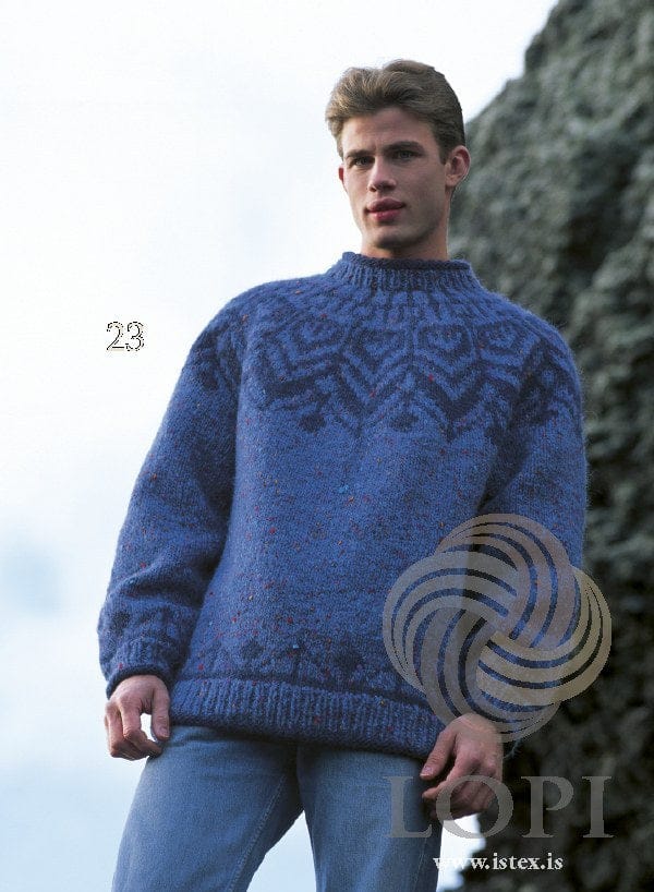 Man by a rock wall in Design 23 of this pattern, shown in the original colours — monet, a mid-blue tweed with small red, pink and turquoise flecks (no longer made), with the yoke and the border above the hem and cuffs in navy, and blue rib. His jeans are not part of the pattern