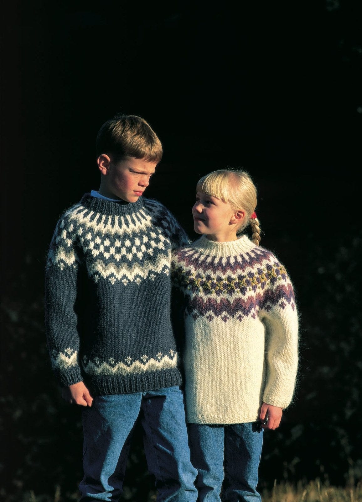 Two children against a dark background in this pattern’s two sweaters, in the original colours. The boy wears Design 24 — slate blue (no longer made) with a yoke of small white diamonds, white and grey zigzags, a band of white diamonds and white spokes, and a matching border above the ribbed hem and cuffs. The girl wears Design 25 — white, with a violet and dusty rose yoke and small embroidered flowers. His shirt collar, her hair tie and their jeans are not part of the pattern