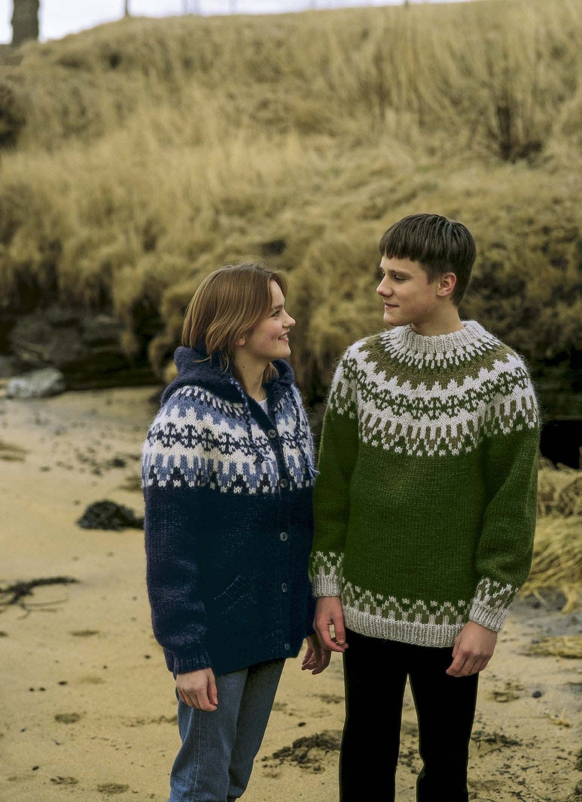 Two young people on a beach. On the right, a young man in the Lopi 18 no. 27 pullover — green, with a pale grey and olive round yoke, a matching border above the pale grey hem and cuff ribs, and a pale grey neckband (shown in the original colours — this kit has garden green, lighter than the green in the photo, light ash heather and highland green in their place). On the left, the hooded blue cardigan from the same leaflet, sold as a separate kit; the trousers are not part of the pattern