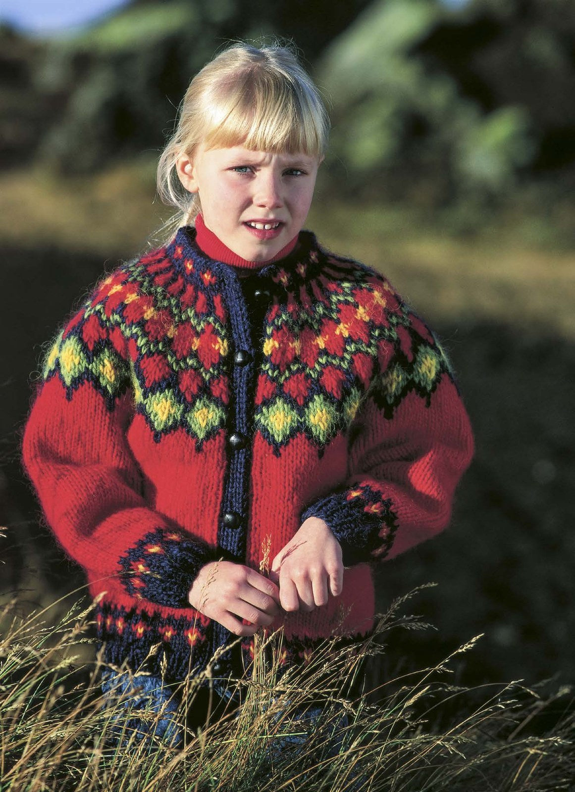 Girl in tall grass in the Lopi 18 no. 31 cardigan, buttoned — bright scarlet with navy front bands, a yoke of green diamonds with yellow centres outlined in navy, red shapes framed in navy and green, navy spokes at the neck, and navy cuffs with small red flowers (shown in the original colours, in strong sun — this kit has a darker happy red, apple green and a deeper, mustard golden heather for 0078, 9212 and 9213). The red polo neck and the jeans are not part of the pattern