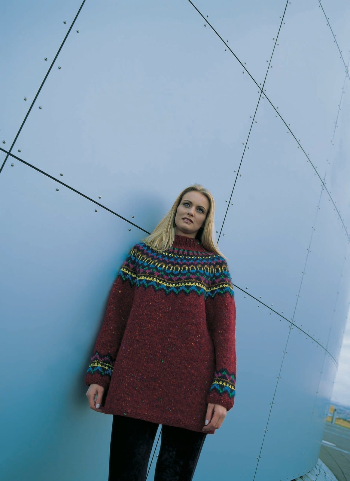 Woman by a blue metal wall in the Lopi 18 no. 34 sweater — long and loose, red tweed with small green, orange and purple flecks, a midnight blue yoke with narrow bands of green, blue, yellow, copper and magenta, the same bands above the moss-stitch cuffs, and a tall red rib collar (shown in the original colours — six are replaced in this kit: a plain ruby red without flecks, a darker green and blue, a deeper yellow, a redder copper and a pinker magenta). The black trousers are not part of the pattern