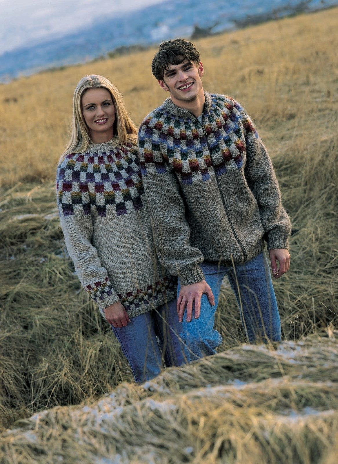 A young man and a woman in long autumn grass. On the right, the man wears the Lopi 18 no. 37 cardigan — oatmeal, zipped, with a checkerboard yoke of small blocks striped blue, purple, dark brown, brick red, gold, teal and dark green beside light beige ones, ribbed welt and cuffs and a high rib collar (shown in the original colours — this kit has five replacements, and its red stripes are a more orange amber). On the left, Design 36, not available as a kit; the T-shirt and jeans are not part of the pattern