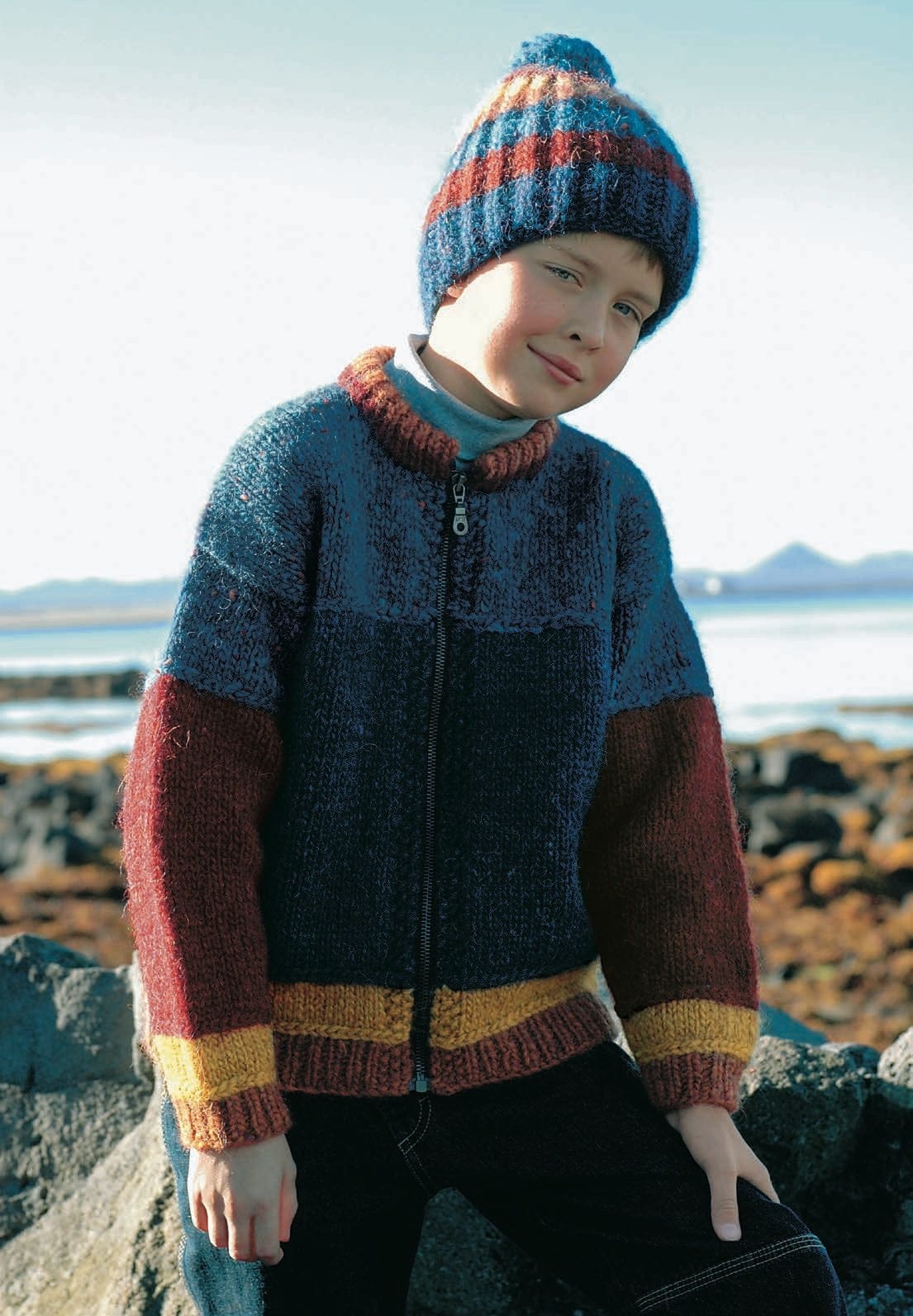Boy on the rocks by the sea in the blue Hlökk zip jacket — a mid-blue tweed yoke and shoulders, a dark navy body, red sleeves, a gold band and amber rib at the hem and cuffs, and a red and amber collar (shown in the original colours — this kit has blue tweed for the monet blue tweed and navy for the sapphire blue). The striped hat is Haddur, a separate kit; the turtleneck and trousers are not part of the pattern