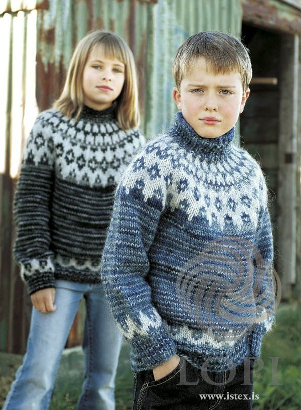 The girl at the back wears the dark grey Ísar sweater — a charcoal body with a light yoke and zigzag bands (shown in the original colour, moon grey, a streaky charcoal yarn — this kit has dark grey heather, an even dark grey, in its place). The boy in front wears the blue version, sold as a separate kit; the jeans are not part of the pattern