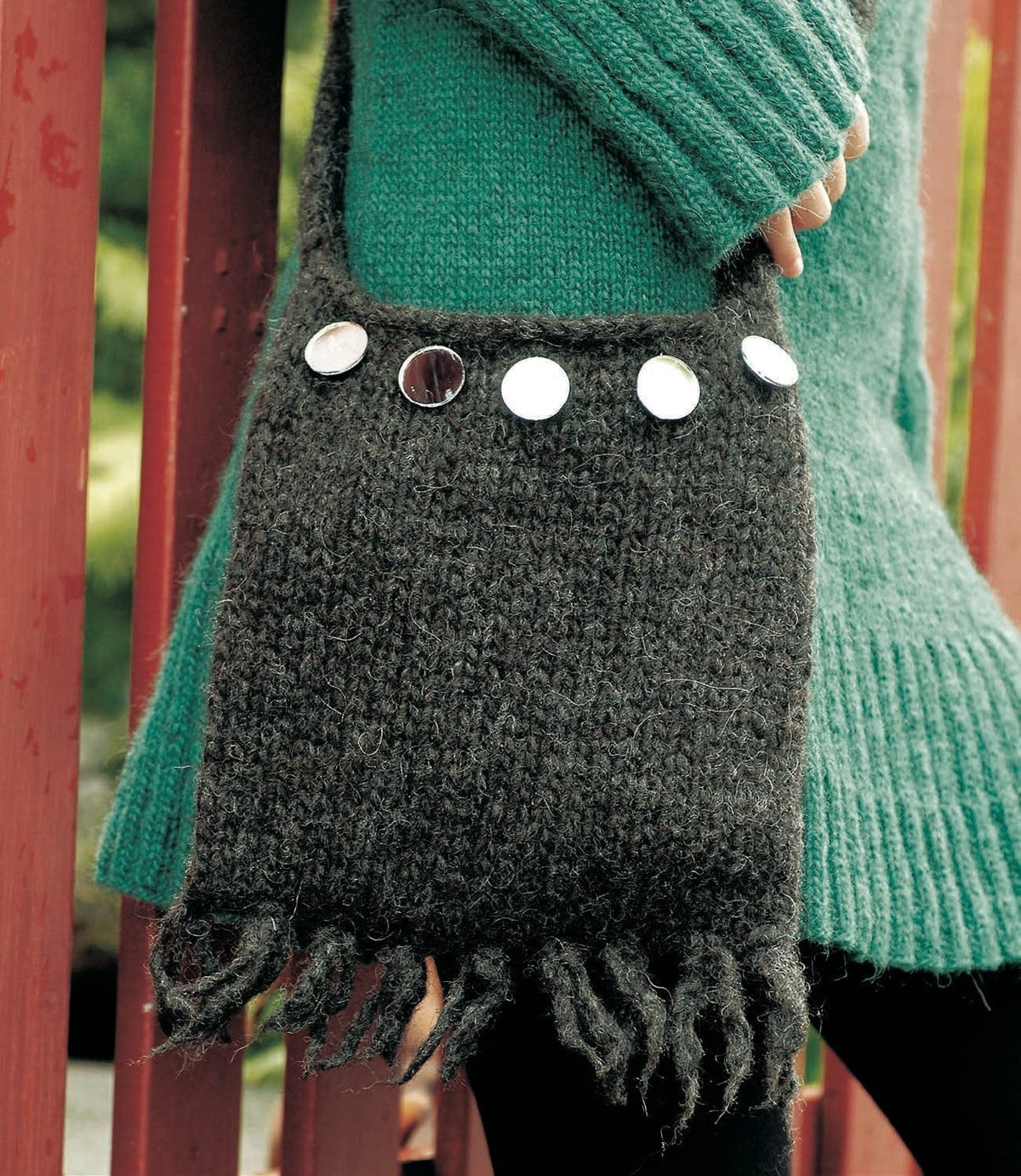The felted Lofn bag worn at the hip, in the pattern’s black heather Jöklalopi — plain stocking stitch, five small round mirror discs sewn in a row across the top, a felted fringe along the bottom and the narrow strap rising from the sides. The long teal sweater is a different pattern