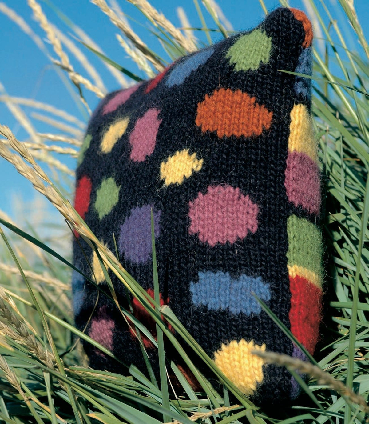 The Opus cushion standing in dune grass — a midnight blue front with big round dots in violet, pink, red, yellow, green, orange and light blue, and the striped back showing at the right-hand edge (shown in the original colours — this kit has space blue, a strong royal blue, for the violet, golden heather for the yellow, amber heather for the orange, winter morning, a paler pink, for the orchid, and light indigo, a greyer blue, for the light blue)