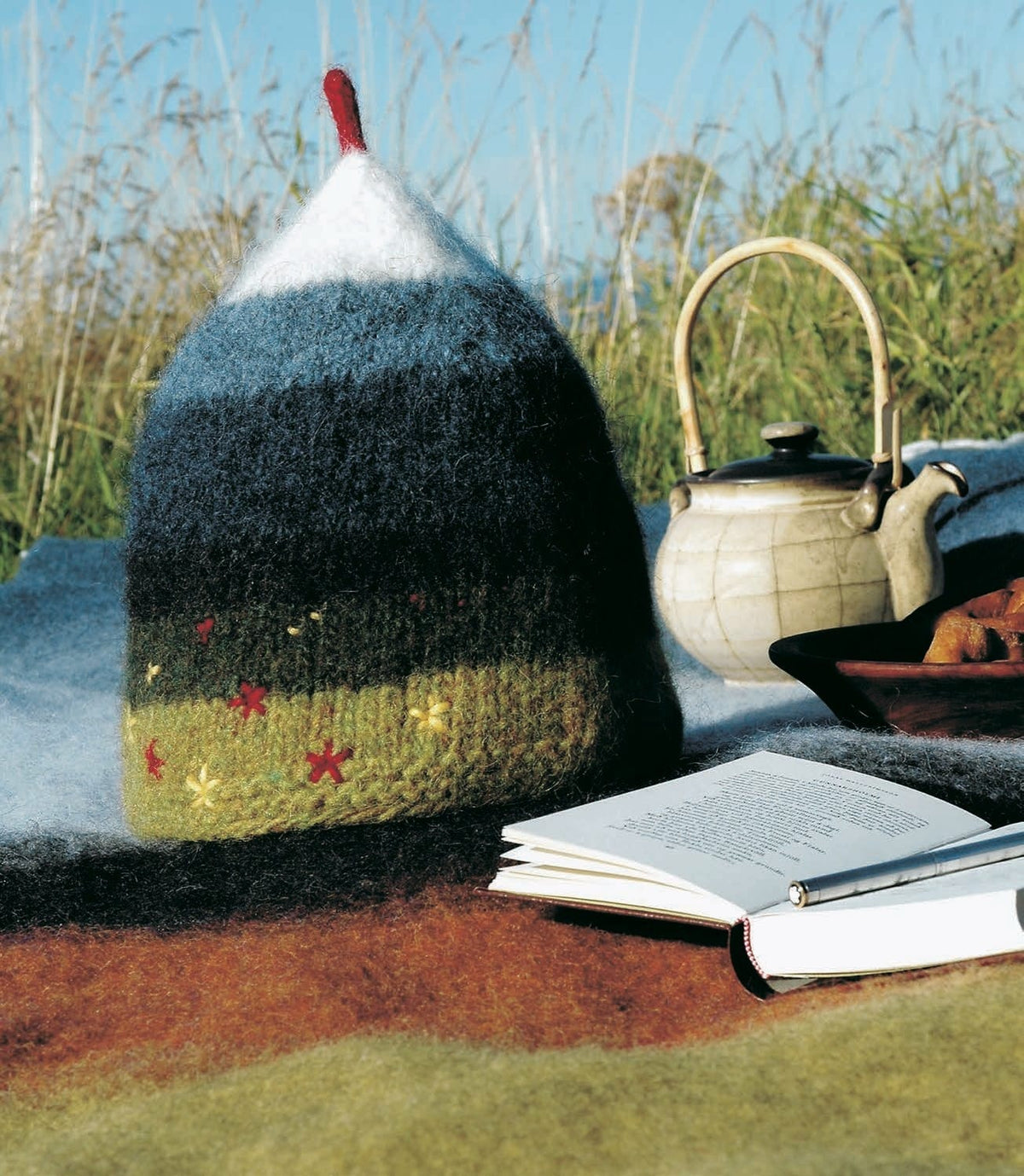 The felted Tindur tea cosy on a wool blanket outdoors — a chartreuse green band and a darker green band with red and yellow embroidered flowers, then black, indigo, light indigo, a white peak and a red tip, shown in the pattern’s original colours. The teapot, the blanket, the bowl, the book and the pen are not part of the pattern