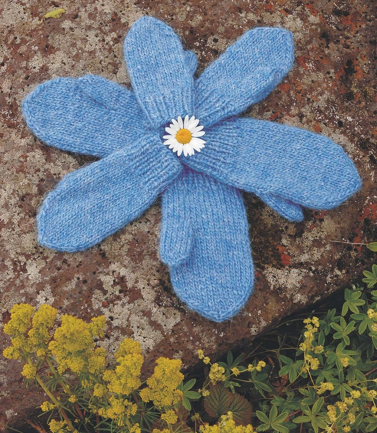 Six Kríli mittens in the pattern’s heaven blue Léttlopi, laid in a flower shape around a daisy on a lichen-speckled rock — plain stocking stitch, K1, P1 rib cuffs, shaped tops and thumbs. The pattern is for one pair