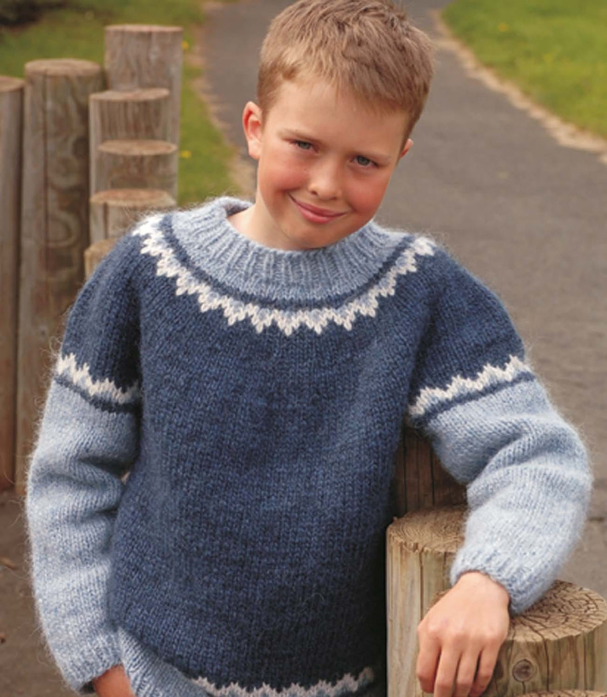 Smiling boy leaning on wooden posts in the T sweater, in the pattern’s colours — an indigo blue body and yoke, light denim sleeves, a white and light denim zigzag band around the upper arms and above the hem, a white zigzag near the top of the yoke and a light denim ribbed neckband