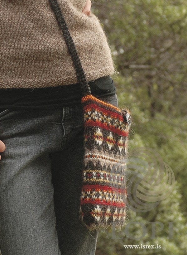 The Taska bag worn across the body on its long, dark garter-stitch strap — bands of diamonds, small crosses and dots in oatmeal, white, yellow, red and orange on a brown-black ground, with an orange top edge and a button, shown in the pattern’s original colours (mustard and orange are no longer made). The light brown sweater, the black top under it and the grey jeans are not part of the pattern