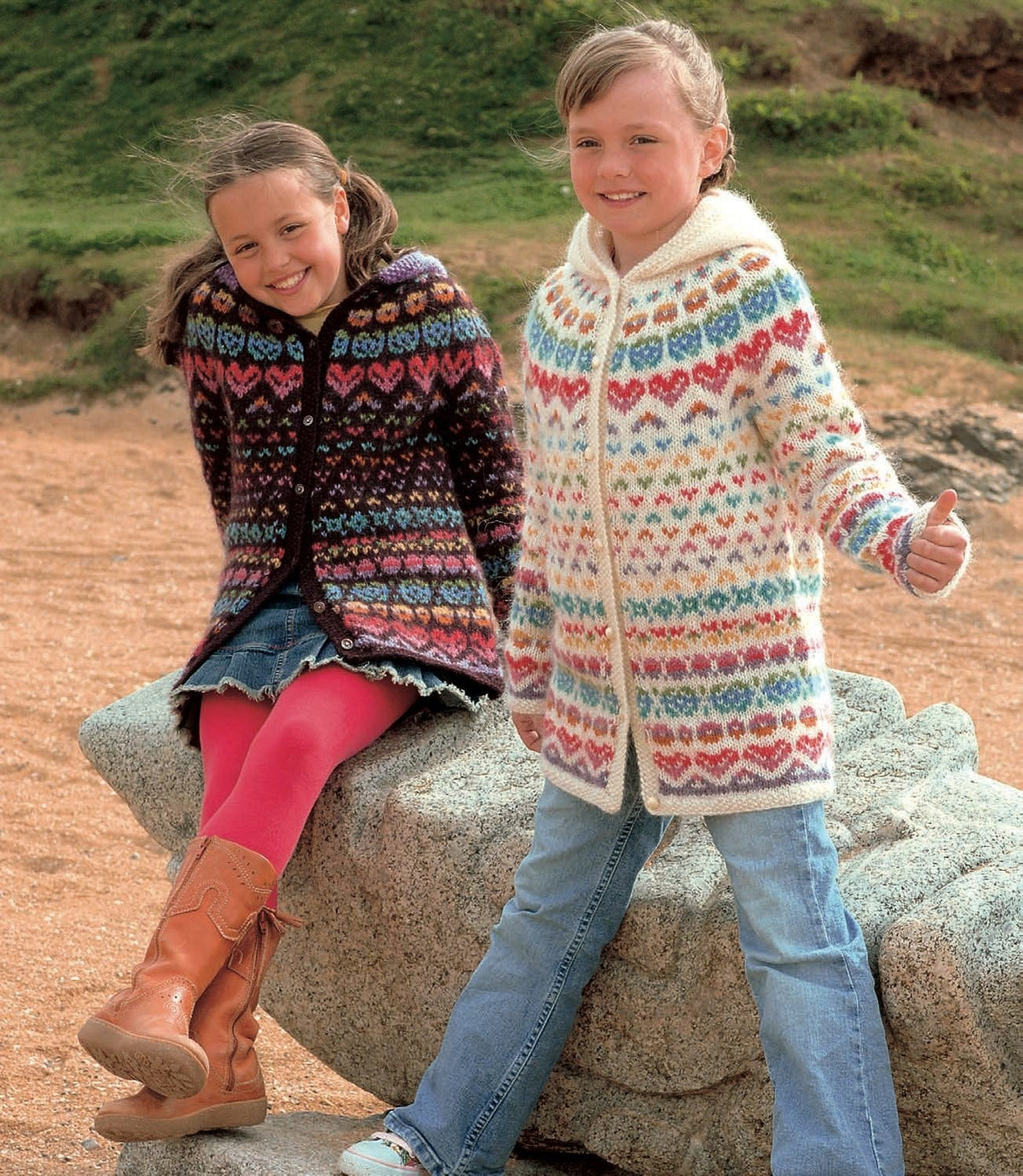 Two girls by a rock on a beach in Ása hooded cardigans, in the pattern’s original colours. Left: the burgundy version — dark wine with rows of bright hearts and small motifs, and a lilac hood. Right: the white version — rows of small motifs in many colours, big red-and-pink hearts round the yoke and above the hem, and a white hood. Dark wine, lilac, light red, orange and sun yellow are no longer made. The yellow top, hair tie, denim skirt, tights, boots, jeans and trainers are not part of the pattern