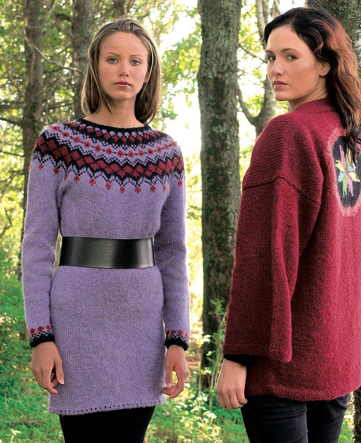 Woman among trees in Eyja, the lilac version, reaching mid-thigh — a lilac dress with a round yoke of black and garnet red zigzags and diamonds, a black neck edge and black cuffs with a garnet border, in the pattern’s original colours (the lilac is no longer made). The wide black belt and the tights are not part of the pattern; the red cardigan and the dark trousers on the woman beside her are not part of it either