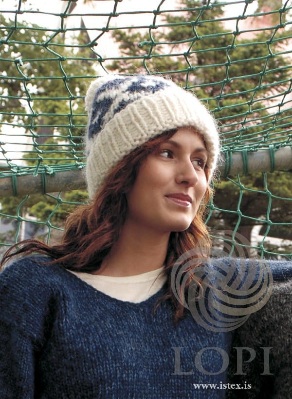 Woman in front of a green goal net wearing Hálka — a white Jöklalopi hat with the deep rib brim turned up and a band of white motifs on mountain blue above it, shown in the pattern’s original colours (mountain blue, 1415, is no longer made). Her blue V-neck sweater, the white T-shirt under it and the dark grey knitted sleeve at the right edge are not part of the pattern