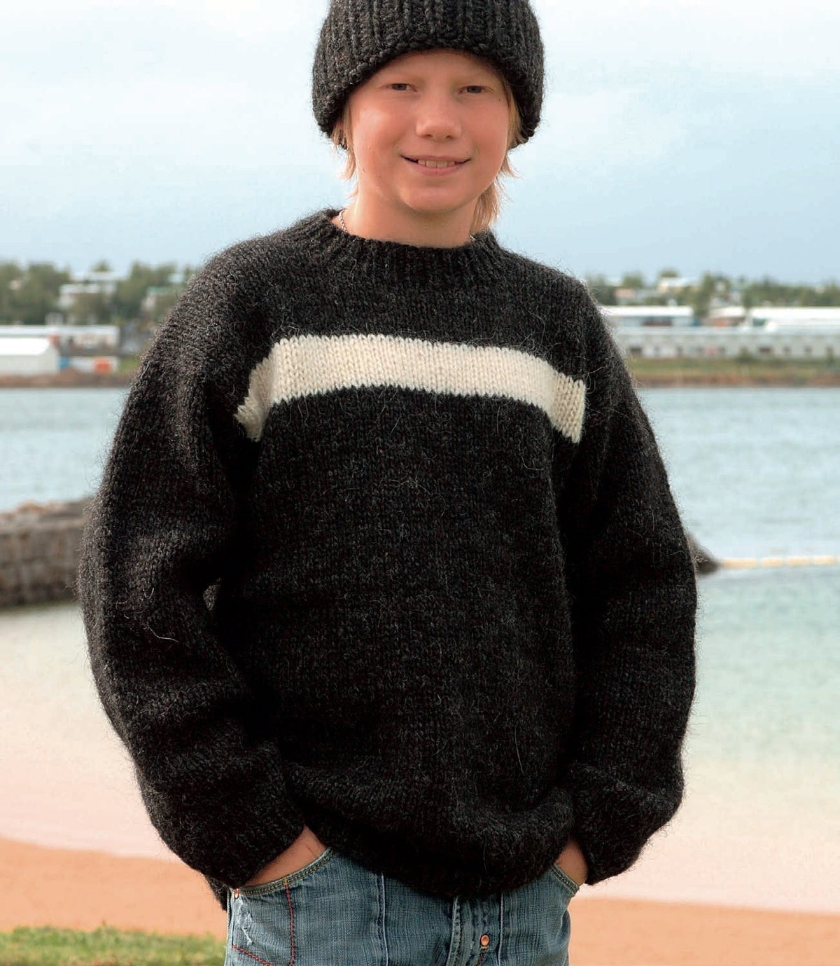 Boy by the sea in Haus, from the front, in the pattern’s colours — a black heather Álafosslopi sweater with one white stripe across the upper chest and a ribbed crew neck. The ribbed hat is Nei, a separate pattern from the same book; the jeans and the necklace are not part of the pattern