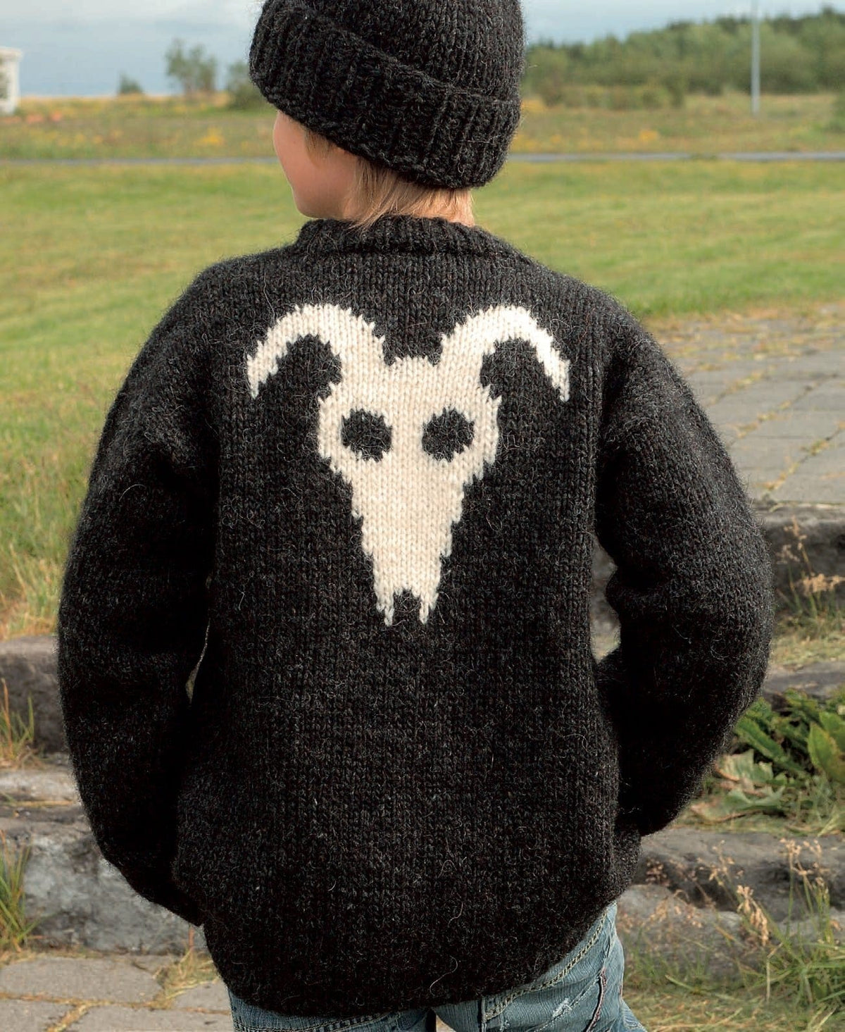 Haus from the back — a big white ram’s head with curved horns and two dark eye holes, knitted in intarsia on black heather. The hat is Nei, a separate pattern; the jeans are not part of the pattern