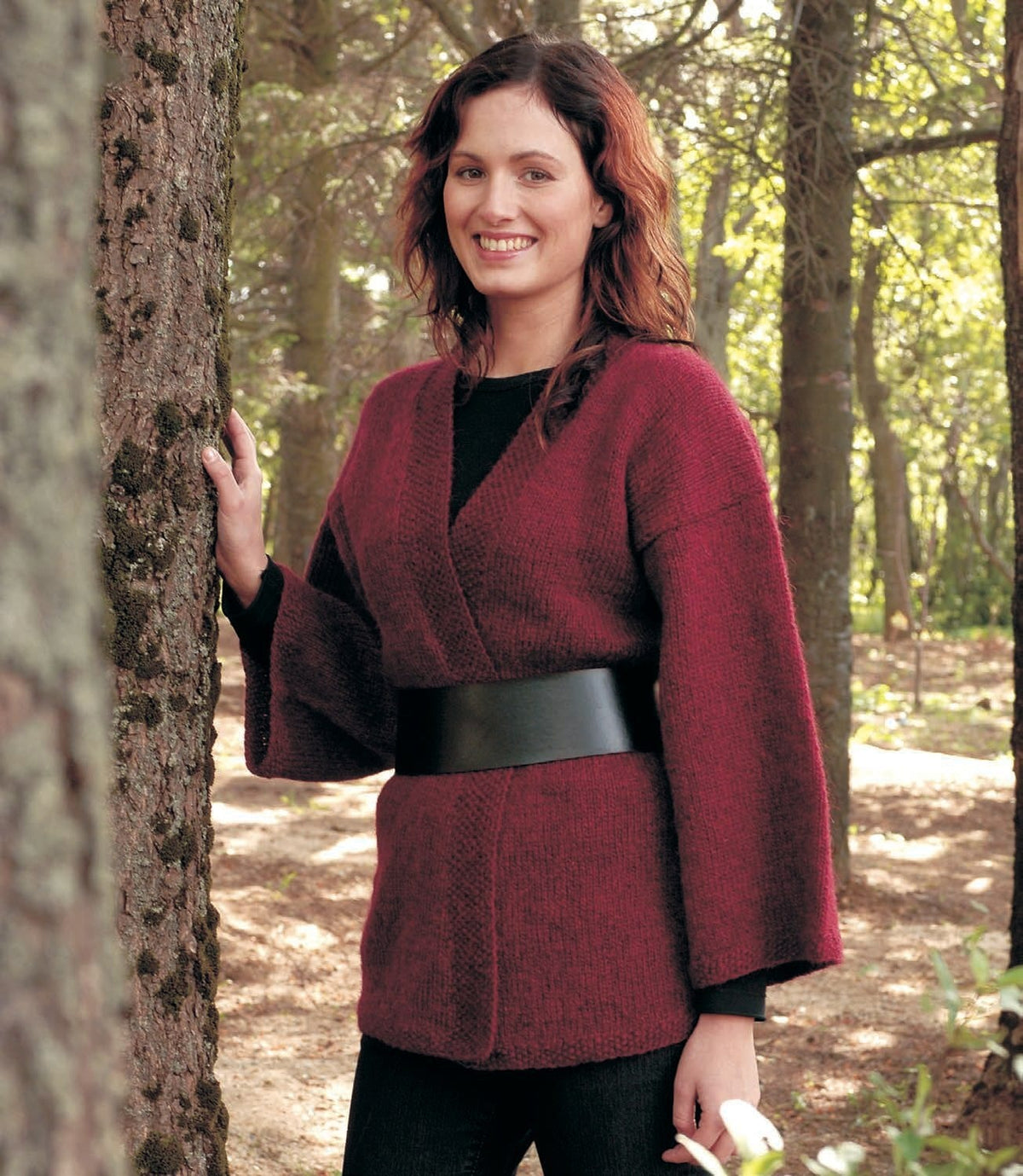 Woman in a wood in Kim, the red version, from the front — garnet red Léttlopi, the fronts crossed over with slanting moss-stitch borders, wide straight sleeves and drop shoulders, shown in the pattern’s own colours. It has no fastening: the wide leather belt is not part of the pattern, nor are the black top and the dark trousers
