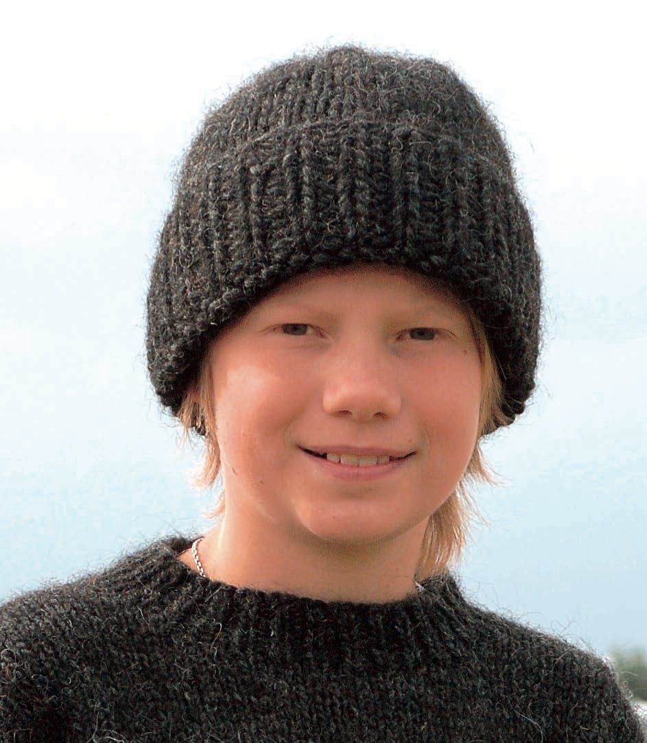 Boy wearing Nei in the pattern’s black heather Jöklalopi — a deep ribbed band, turned up in the photo, and a plain stocking-stitch crown. The sweater is Haus, a separate pattern from the same book; the necklace is not part of the pattern