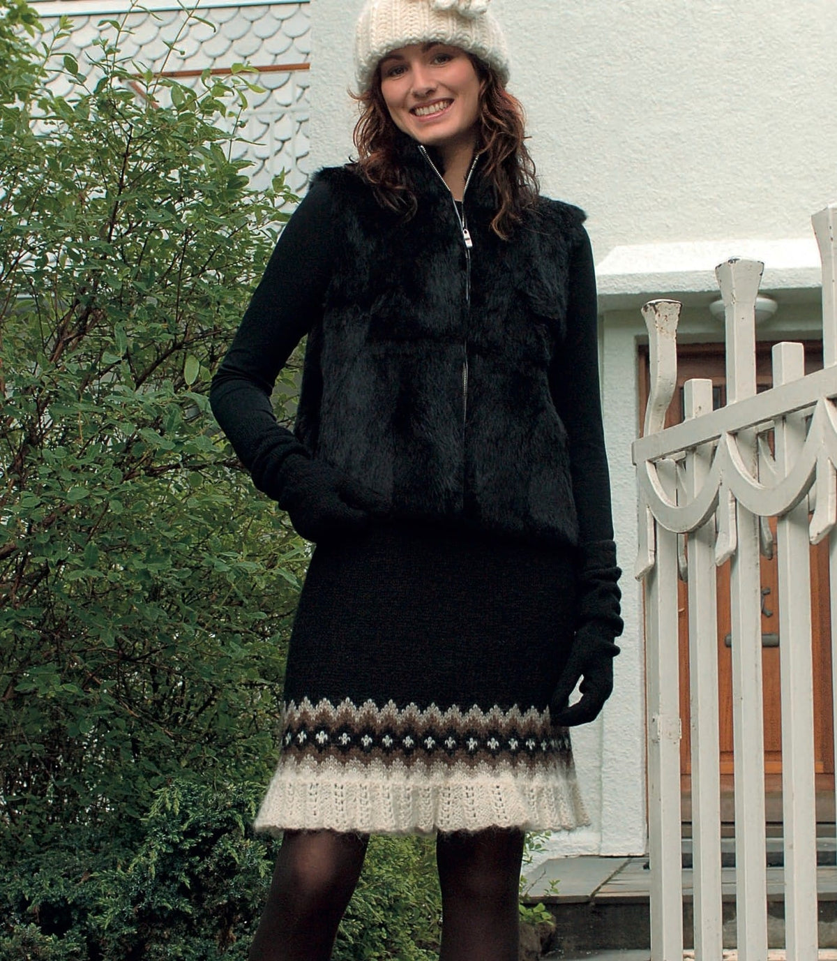 Smiling woman by a white wooden gate in Pífa, shown in the pattern’s own colours — a black flared skirt ending just above the knee, with a border of light beige and acorn zigzags around a black band of small white crosses, and a white eyelet-lace frill at the hem. Her white hat with a crocheted flower is Snjór, a different pattern from the same book; the black fur vest, black top, long black gloves and dark tights are not part of the pattern