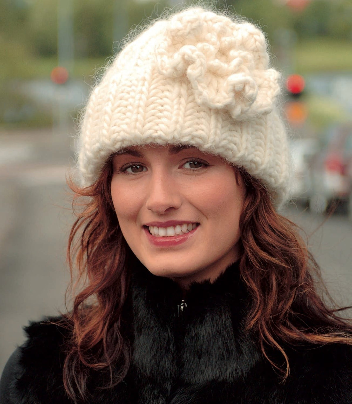Smiling woman in Snjór, shown in the pattern’s white Jöklalopi — a chunky brioche-stitch hat with deep ribs, a turned-up brim and a large crocheted flower with chain loops at its centre, near the top at one side. Her black fur jacket is not part of the pattern