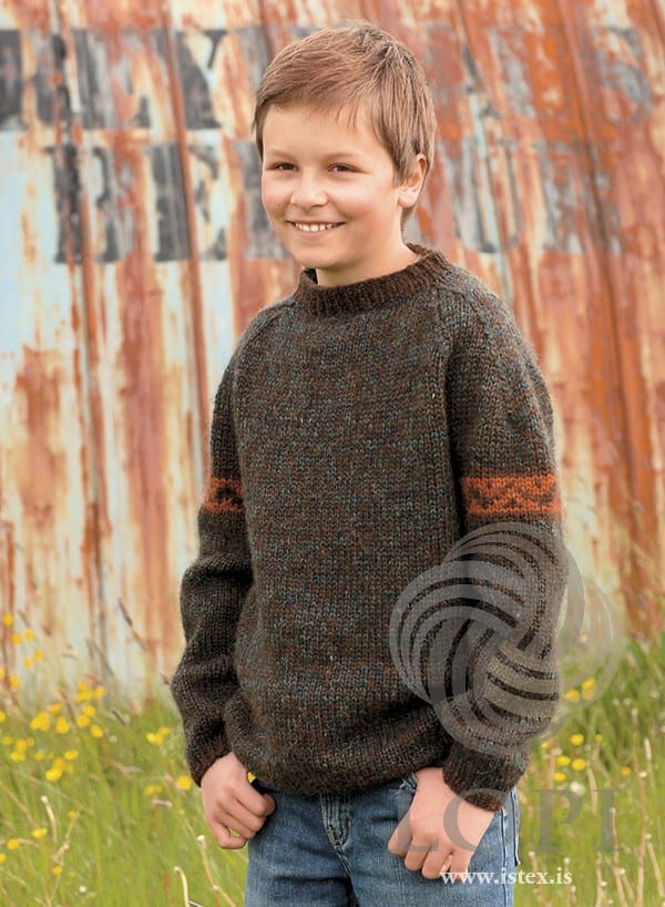 Smiling boy by a rusty corrugated-iron wall in Tat — a brown marl crew-neck sweater flecked with grey-teal, a dark brown rib neckband and a dark amber zigzag band on each upper sleeve, shown in the pattern’s original colours (the sea green in the marl is no longer made). His jeans are not part of the pattern