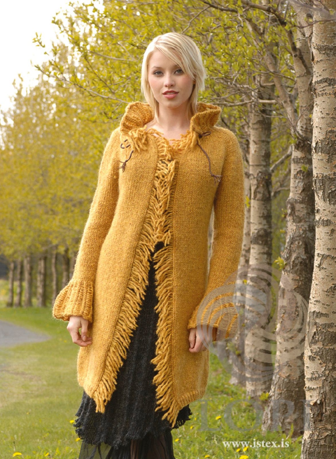 Woman among trees in Drama, the yellow coat — a long open coat in golden (9964) Álafosslopi with a fringe down both fronts, ruffled cuffs, a tall ruffled collar and a thin brown cord at the neckline, shown in the pattern’s own colour. The black skirt is not part of the pattern