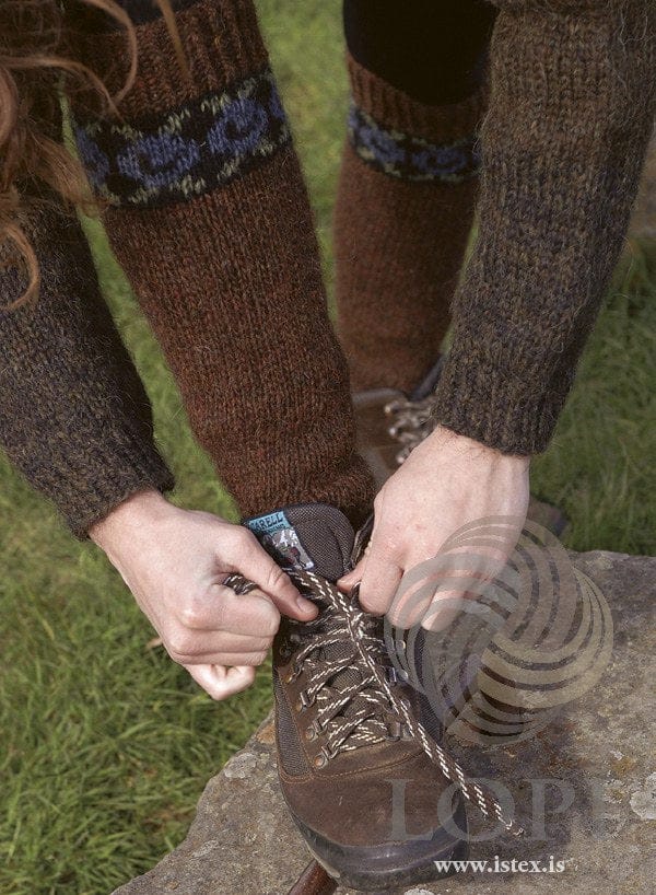 Leggur leg warmers in the Roses version, worn while lacing a hiking boot on a rock — hazel brown with a twisted-rib top and a band of stone blue roses and celery green leaves on black just below it, shown in the pattern’s original colours (hazel is no longer made). The brown sweater, the dark trousers and the hiking boots are not part of the pattern
