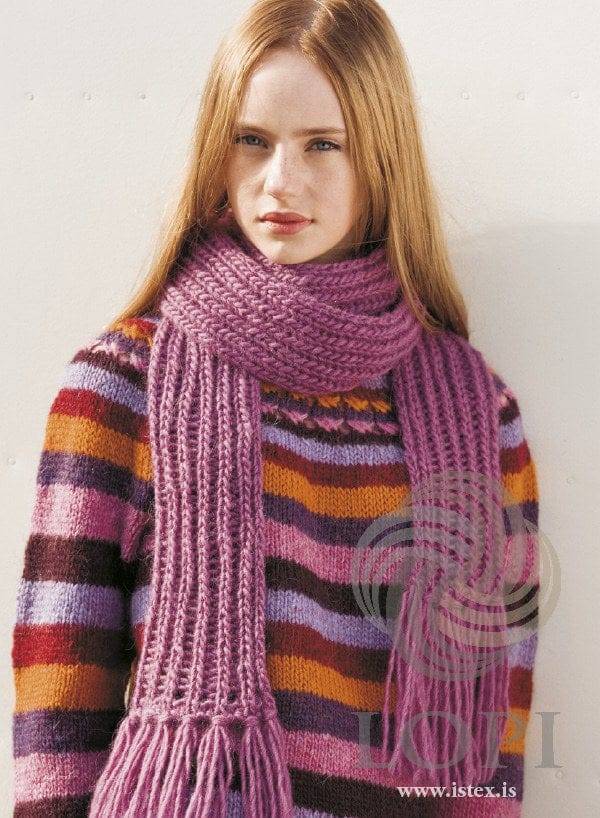 Young woman wearing Glypja, shown in the pattern’s original orchid (0159, no longer made) — a dusty pink-purple Álafosslopi scarf wrapped once round her neck, with open ladders between the ribs and a tassel fringe at both ends hanging down the front. Her striped sweater is a different pattern (Kross, Lopi 28) and is not part of this pattern