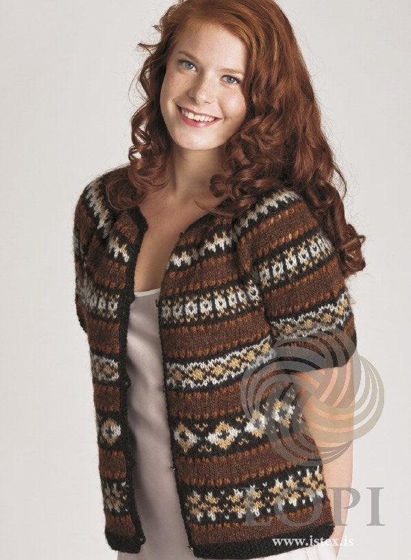Woman in the Hún cardigan, worn open — short sleeves, bands of stars, diamonds and crosses, brown stripes, rust eyelet rows, black moss-stitch edges and button loops (shown in the original colours — this kit has chocolate heather for the hazel and barley for the camel). The satin top under it is not part of the pattern