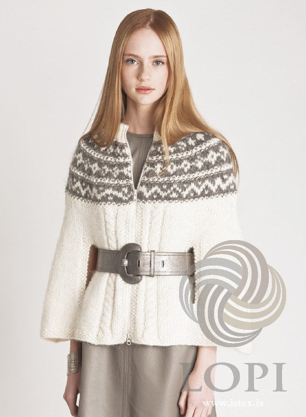 Woman in the white Rósn cape, zipped to the neck — cables, moss-stitch edges and a yoke in white and grey heather. The wide belt and the dress are not part of the pattern