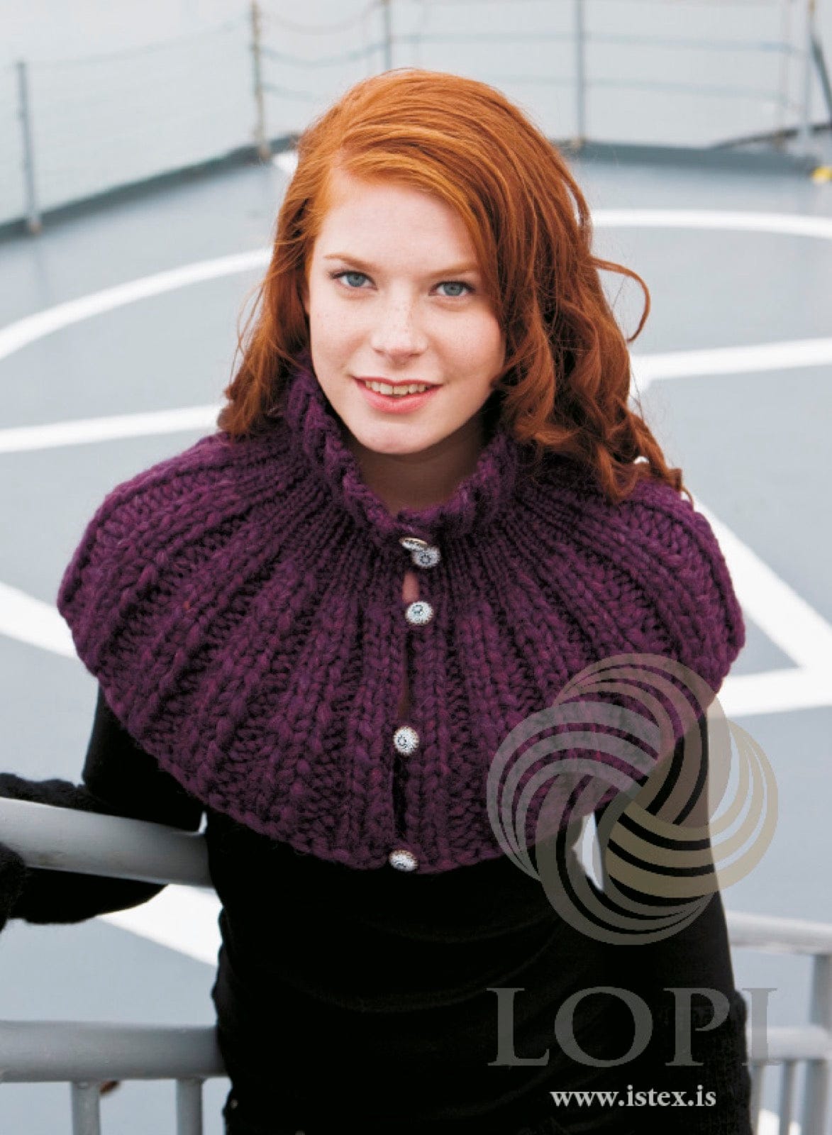 Woman on a ship’s deck in Rú — a plum heather Plötulopi shoulder collar in rib, standing up at the neck and flaring over the shoulders, with round silver-tone buttons down the front, shown in the pattern’s own colour. The black top is not part of the pattern