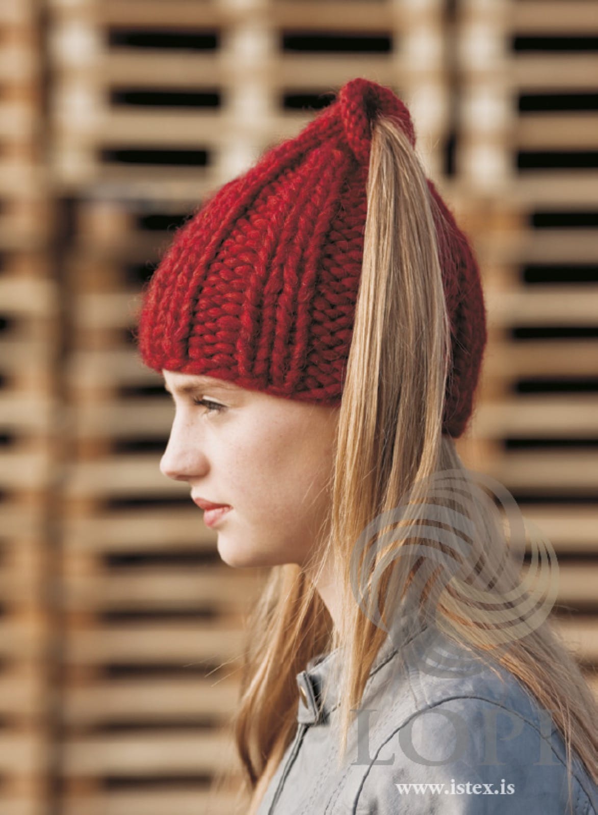 Young woman in profile in Strompur — a carmine red Plötulopi hat in chunky rib, her ponytail coming out through the open, rolled top, shown in the pattern’s own colour. The grey jacket is not part of the pattern