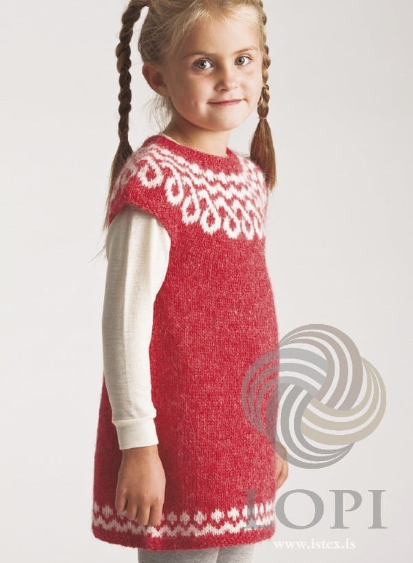 Girl with long braids in Teikn, the red version — a round yoke of white figures that read as interlocking loops, little cap sleeves, an A-line skirt and a white band above the moss-stitch hem, shown in the pattern’s original light red (no longer made). The cream long-sleeved top, the grey leggings and the hair ties are not part of the pattern