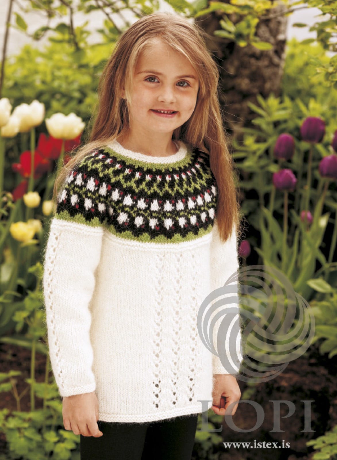 Girl in front of tulips wearing Bláklukka, the white version — a white body with an eyelet panel up the front and up each sleeve, garter ridges at the hem and at the base of the yoke, and a black yoke band with white bell flowers, small crimson dots and green zigzags, shown in the pattern’s original colours. The black leggings are not part of the pattern