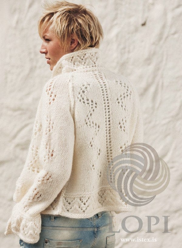 Woman in the white Elding cardigan, seen from the back — a column of eyelets down the centre back with a zigzag lace line on each side, the raglan seams, a lace border at the hem, a flared lace cuff and the collar standing up at the neck. The jeans are not part of the pattern