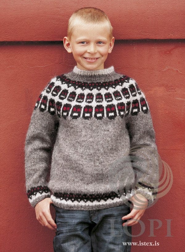 Smiling boy against a red wall in Fés, the grey version, in the pattern’s own colours — a grey Léttlopi sweater with three rows of black faces with crimson eyes and white mouths on ash in the yoke, and a matching dotted border above the rib at the hem and cuffs. His jeans are not part of the pattern