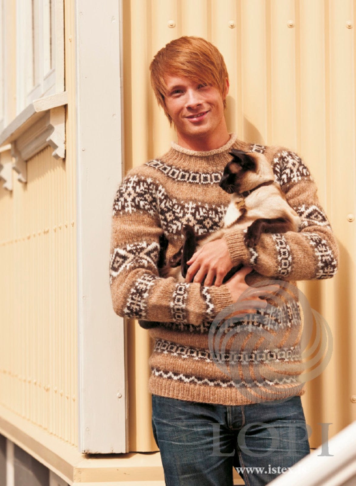 Man holding a Siamese cat, wearing Hrím in the beige colourway — a barley body and sleeves with bands of zigzags, squares, crosses and diamonds in browns and white, a patterned yoke and a straw edge rolled over at the neck, shown in the pattern’s original colours. The jeans and the cat are not part of the pattern