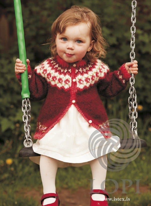 Toddler on a swing in Knúpur, the red version — garnet red with a white yoke band of bright red flowers and green dots, bright red picot edges and front border, flower borders above the hem and cuffs, and white buttons, shown in the pattern’s original colours (light red is no longer made). The white dress, white tights and red shoes are not part of the pattern