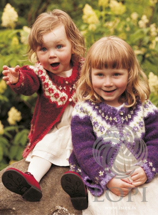 Two girls in Knúpur cardigans — the red version on the left (shown in the original colours — this kit has crimson red in place of the light red) and the purple version on the right, sold as a separate kit. The white dresses, the tights and the red shoes are not part of the pattern