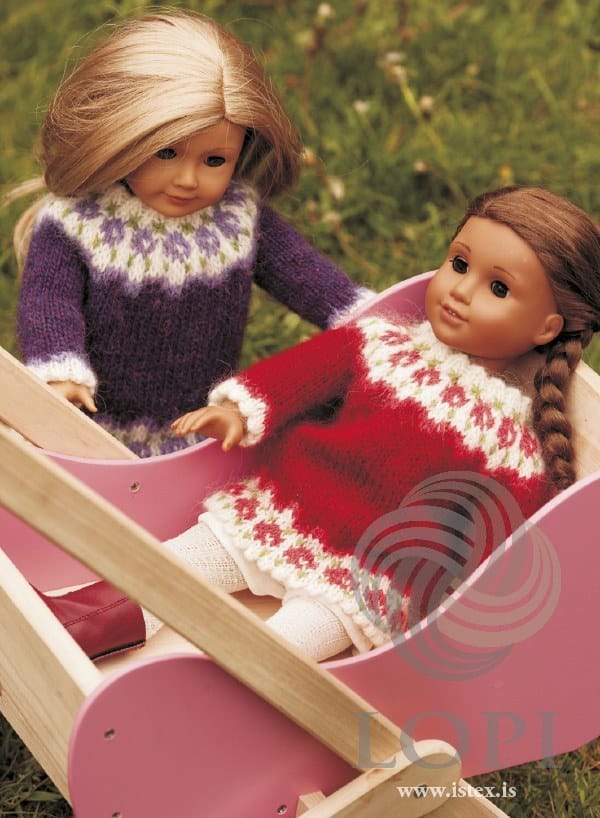 Two dolls in a pink toy cradle: at the back, the purple Rós doll sweater (original lilac roses — this kit has milkyway in their place); in front, the red version, sold as a separate kit. The cradle and the white tights are not part of the pattern