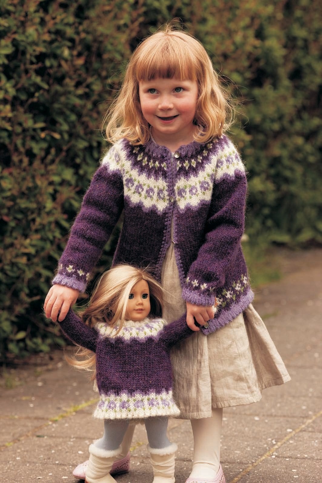 A girl holds the hands of a doll in the purple Rós doll sweater — violet, with white bands of lilac roses and green dots at the hem and yoke, and white picot edges (shown in the original colours — this kit has milkyway, a paler, bluer lavender, for the lilac). The girl wears the matching Knúpur cardigan, sold as a separate kit; her dress and the doll’s tights and boots are not part of the pattern