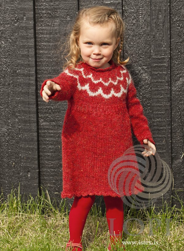 Little girl in front of a black wooden wall in Bára — a carmine red Plötulopi dress with two bands of ash scallop shapes on the round yoke and a crocheted shell edging at the hem, cuffs and neck, in the pattern’s original colours. The small hair tie in her braid, her red tights and red shoes are not part of the pattern