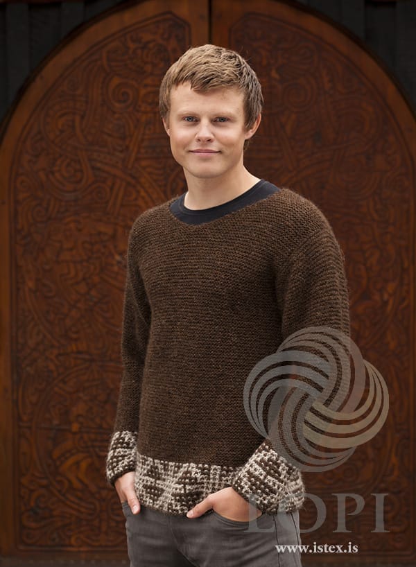 Young man in front of a carved wooden door in Örk — chocolate Léttlopi in garter stitch with light beige and chocolate slip-stitch borders at the hem and cuffs, in the pattern’s original colours. The dark T-shirt under it and his grey trousers are not part of the pattern
