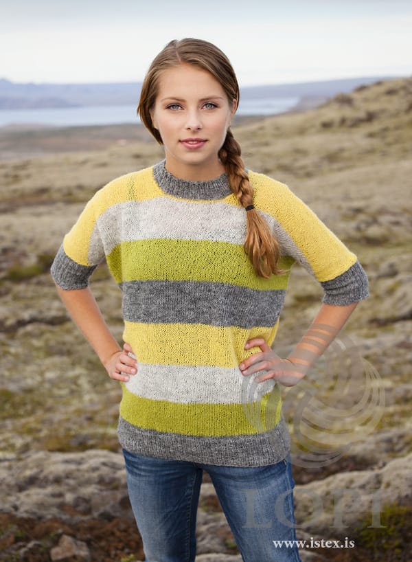 Young woman with a long side braid, hands on hips, on a moorland hillside in Blaka — a wide Einband sweater in broad stripes of lime, ash heather (pale grey), yellow and grey heather, yellow across the shoulders, with grey ribbed cuffs ending at the elbow, a grey hem rib and a grey neckband, shown in the pattern’s own colours. The blue jeans and the hair elastic are not part of the pattern