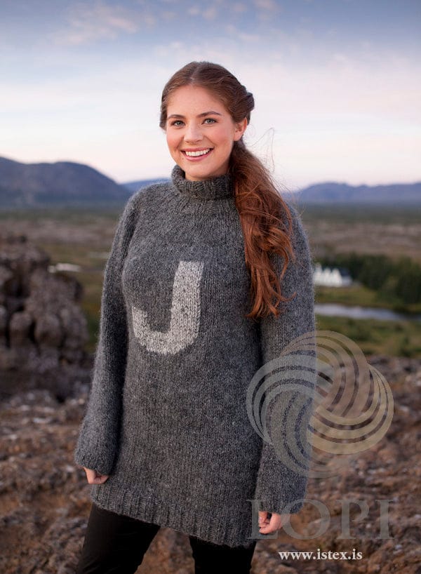 Woman outdoors in the long ladies’ Frikki, shown in the pattern’s own colours — dark grey Álafosslopi with a tall ribbed collar folded over and a light ash J sewn on the front. The black trousers are not part of the pattern