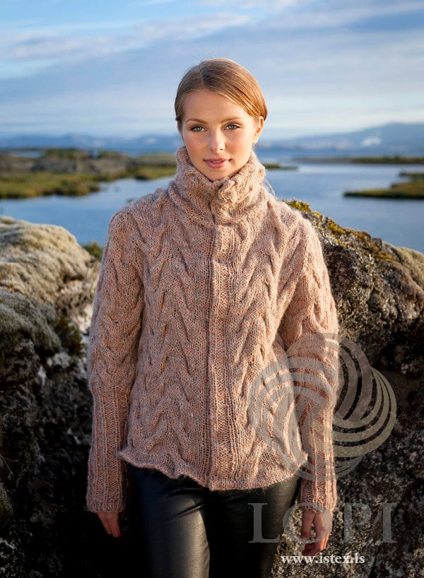 Woman standing among mossy lava rocks by a lake in Kaðlastaðir — a cardigan cabled all over in a blended pinkish beige, worn closed, with a tall folded cabled collar, a narrow front border and ribbed lower sleeves, shown in the pattern’s own colours (peach Einband and light beige Plötulopi held together). The black leather-look trousers are not part of the pattern
